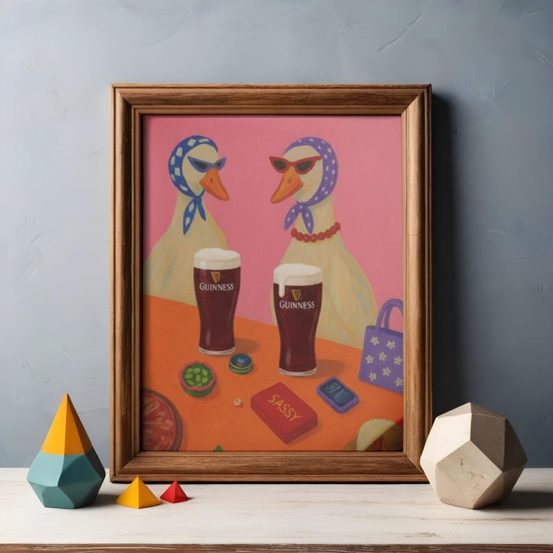 K&ouml;nnte beinhalten: Gerahmter Kunstdruck mit zwei G&auml;nsen, die Sonnenbrillen und Kopft&uuml;cher tragen und Guinness genie&szlig;en. Das Kunstwerk hat einen rosa Hintergrund und einen Holzrahmen. Eine kleine rote Schachtel mit dem Wort "SASSY" ist sichtbar.