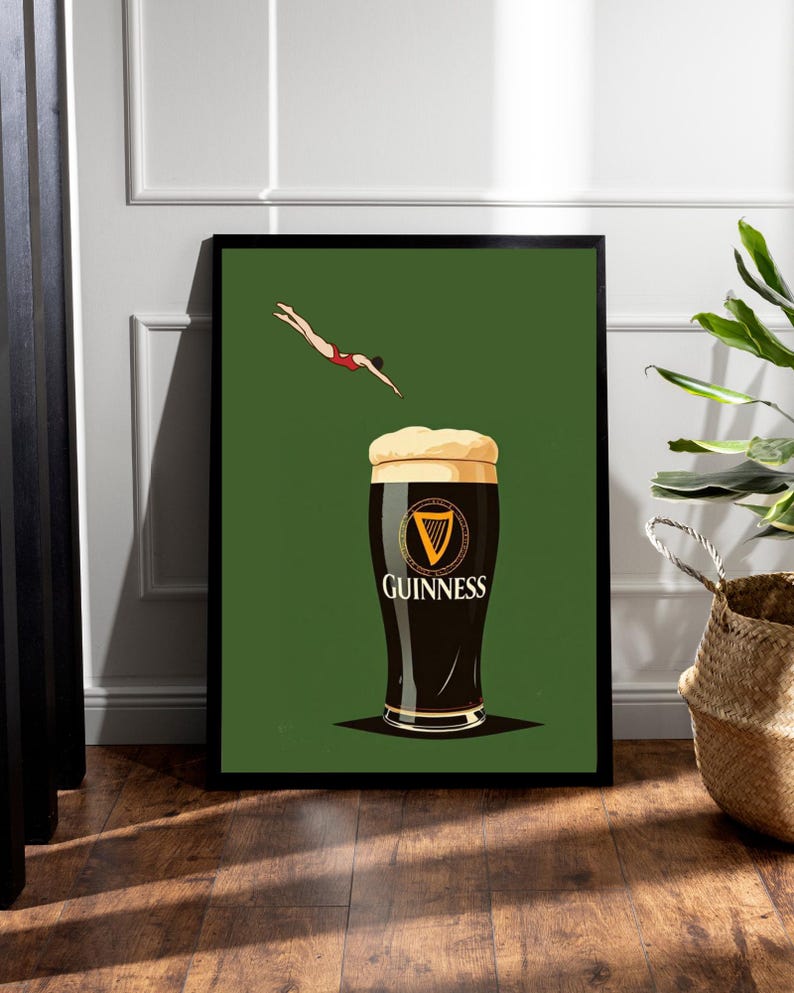 May include: A framed art print featuring a pint of Guinness with a diver in a red swimsuit. The Guinness glass is black with a golden rim and the Guinness logo. The background is green.