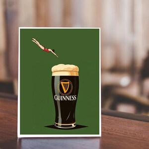 May include: A print featuring a pint of Guinness stout with a diver in a red swimsuit. The Guinness logo is prominently displayed on the glass against a green background. The image is set on a wooden surface.
