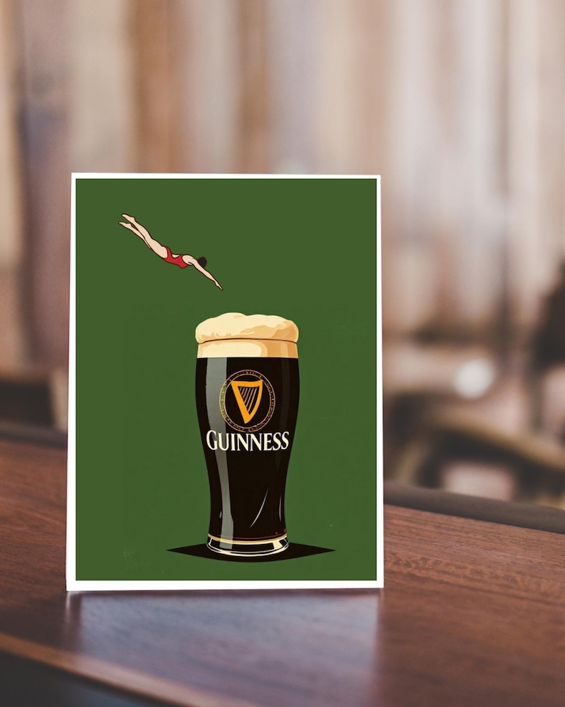 May include: A print featuring a pint of Guinness stout with a diver in a red swimsuit. The Guinness logo is prominently displayed on the glass against a green background. The image is set on a wooden surface.