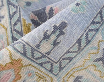 May include: Close-up of a rolled-up area rug with a detailed, geometric pattern. The rug features a light blue background with accents of teal, pink, and gold. The design includes stylized floral and geometric shapes, creating a vintage aesthetic.
