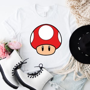 Puede incluir: Camiseta blanca con un gr&aacute;fico de hongo rojo con lunares blancos, contorno negro y tallo beige. La camiseta se muestra con botas blancas, un sombrero negro y una bufanda color crema. El dise&ntilde;o recuerda a un videojuego popular.