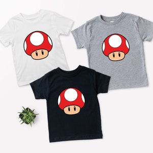 Puede incluir: Tres camisetas de manga corta en blanco, gris y negro, cada una con un dise&ntilde;o de hongo de dibujos animados. El hongo tiene un gorro rojo con lunares blancos, un tallo marr&oacute;n y ojos negros. Las camisetas son para ni&ntilde;os.
