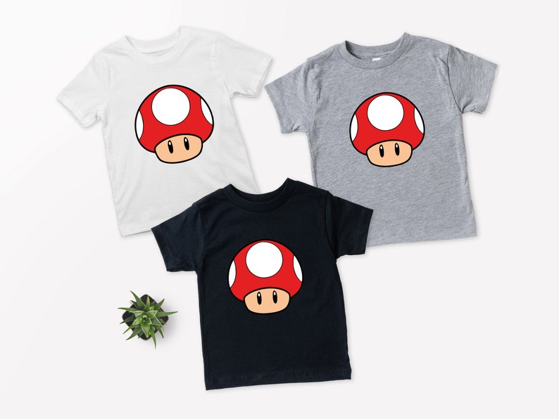 Puede incluir: Tres camisetas de manga corta en blanco, gris y negro, cada una con un dise&ntilde;o de hongo de dibujos animados. El hongo tiene un gorro rojo con lunares blancos, un tallo marr&oacute;n y ojos negros. Las camisetas son para ni&ntilde;os.