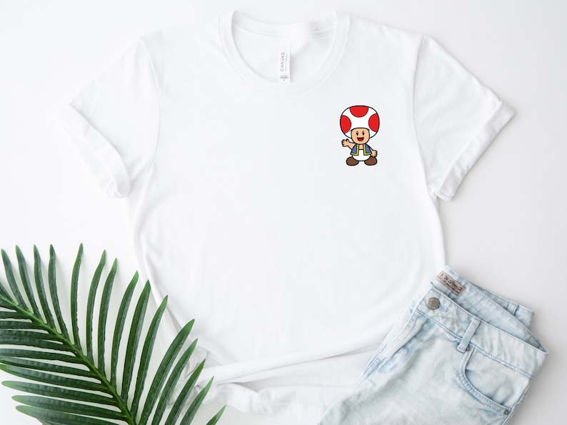 May include: White t-shirt with a cartoon image of a red-capped mushroom character with a blue vest and brown pants. The character is waving with its right hand.