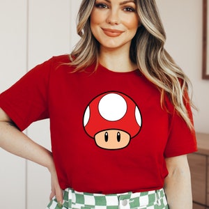 Puede incluir: Camiseta roja con un dise&ntilde;o de hongo de dibujos animados. El hongo tiene una gorra roja con manchas blancas, un tallo beige y ojos negros. La camiseta es de color rojo liso.