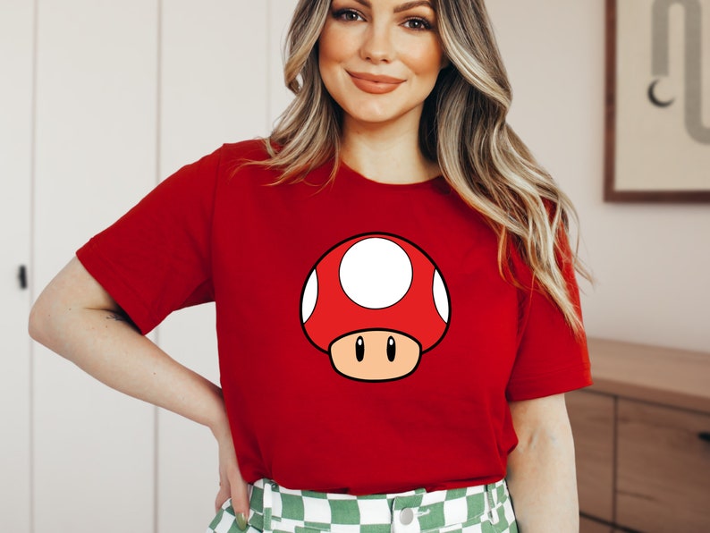 Puede incluir: Camiseta roja con un dise&ntilde;o de hongo de dibujos animados. El hongo tiene una gorra roja con manchas blancas, un tallo beige y ojos negros. La camiseta es de color rojo liso.