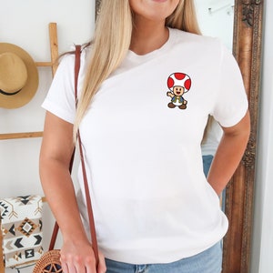 May include: White t-shirt with a small embroidered image of a cartoon mushroom character wearing a blue shirt and a red cap.