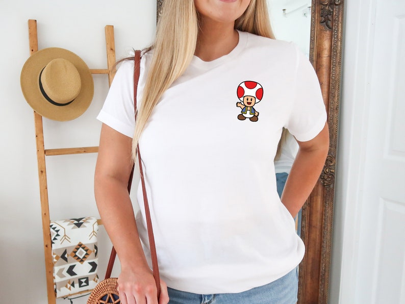 May include: White t-shirt with a small embroidered image of a cartoon mushroom character wearing a blue shirt and a red cap.