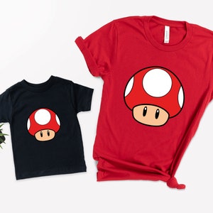 Puede incluir: Dos camisetas de manga corta, una negra y otra roja, cada una con un dise&ntilde;o de hongo de dibujos animados. El hongo tiene un sombrero rojo con lunares blancos, un tallo marr&oacute;n y ojos negros. Las camisetas son probablemente para ni&ntilde;os o adultos.