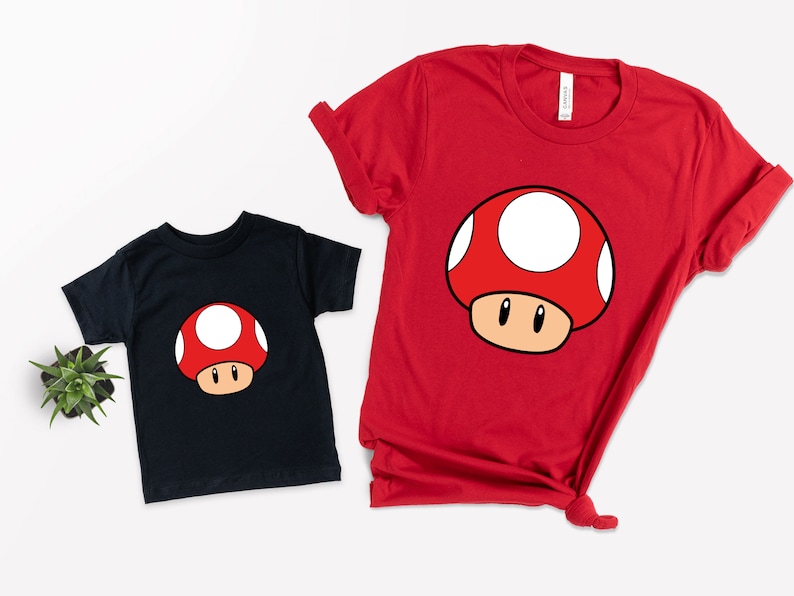 Puede incluir: Dos camisetas de manga corta, una negra y otra roja, cada una con un dise&ntilde;o de hongo de dibujos animados. El hongo tiene un sombrero rojo con lunares blancos, un tallo marr&oacute;n y ojos negros. Las camisetas son probablemente para ni&ntilde;os o adultos.