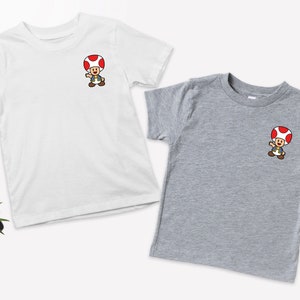 May include: Two t-shirts with a cartoon image of a mushroom character. The shirt on the left is white and the shirt on the right is gray.
