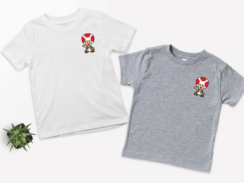 May include: Two t-shirts with a cartoon image of a mushroom character. The shirt on the left is white and the shirt on the right is gray.
