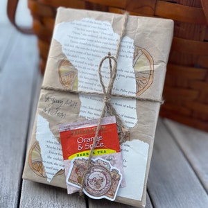 May include: A wrapped book with torn paper edges, tied with twine, and adorned with a tea bag and a camera sticker. The tea bag reads "Orange & Spice Herbal Tea." The book is labeled "Young Adult Fiction."