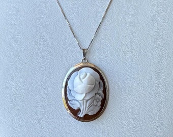 Pode incluir: Um colar de corrente de prata com um pingente de camafeu oval que apresenta uma rosa branca esculpida em um fundo marrom.
