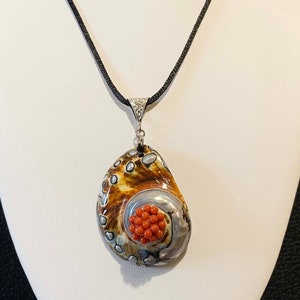 Op de afbeelding: Een zilveren ketting met een hanger van een bruine en grijze schelp met een cluster van rode koraal kralen. De ketting is aan een zwart koord.