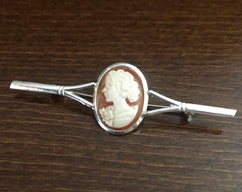 Pode incluir: Um broche de prata com um camafeu de perfil de mulher em branco sobre fundo marrom.
