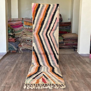 Puede incluir: Una alfombra larga y estrecha con un patrón de rayas coloridas en tonos de rosa, naranja, negro y blanco. La alfombra tiene flecos en el borde inferior.