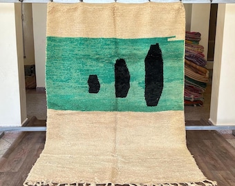 May include: A rectangular rug with a beige background and a central turquoise stripe. Three black abstract shapes are on the turquoise stripe. The rug has a fringe along the bottom edge. The rug is displayed on a metal rack.