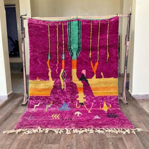 Peut inclure: Un tapis marocain rose vif avec un motif géométrique mettant en valeur un motif central vert et orange. Le tapis a une bordure à franges et est présenté sur un support en bois.