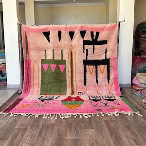 May include: A large rectangular area rug featuring a light pink background with a variety of geometric shapes and abstract symbols in black, olive green, and bright pink. The rug has a thick fringe along the bottom edge and a woven texture.