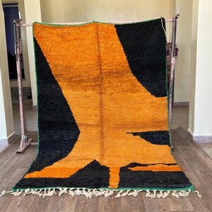May include: A large, rectangular rug with a bold geometric design. The rug features a striking contrast of black and orange, with a green border and white fringe. The design is abstract, with large shapes dominating the surface. The rug is displayed on a metal rack.
