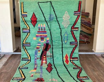 May include: A green rug with a geometric pattern. The rug has a central design with red, yellow, blue, and black accents. It has a fringe along the bottom edge and is displayed on a metal rack.
