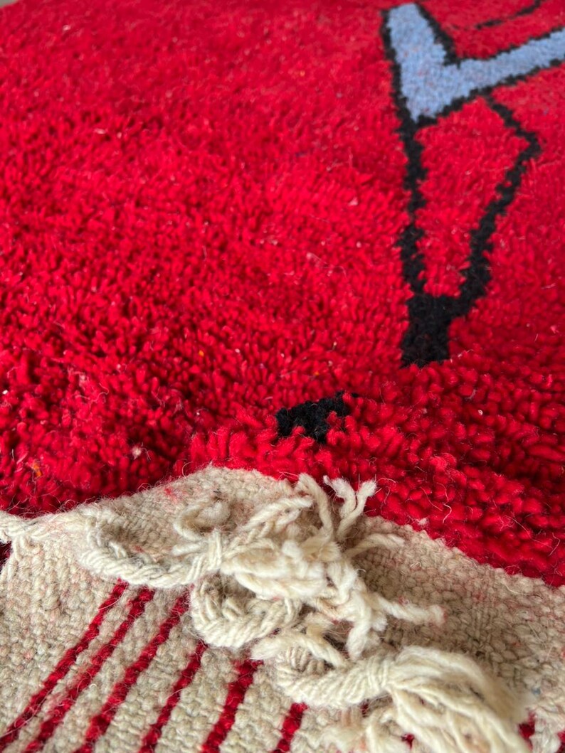 Peut inclure: Un tapis rouge &eacute;pais &agrave; poils longs pr&eacute;sentant un motif graphique noir et bleu clair. Le tapis poss&egrave;de un bord tiss&eacute; beige visible avec des rayures verticales rouges et des pompons &agrave; franges nou&eacute;s d&eacute;coratifs sur le bas de la pi&egrave;ce textile.