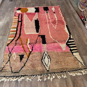 May include: A pink, brown, and black Moroccan rug with a geometric pattern. The rug has a fringe border.