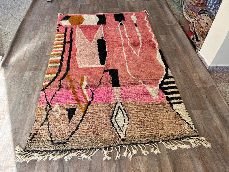 May include: A pink, brown, and black Moroccan rug with a geometric pattern. The rug has a fringe border.