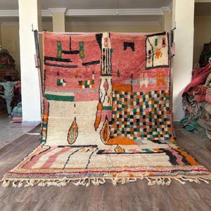 Op de afbeelding: Een groot, kleurrijk tapijt met een geometrisch ontwerp. Het tapijt heeft een mix van kleuren, waaronder roze, oranje, groen en wit. Het tapijt heeft een franje langs de onderrand en wordt gepresenteerd op een houten vloer.