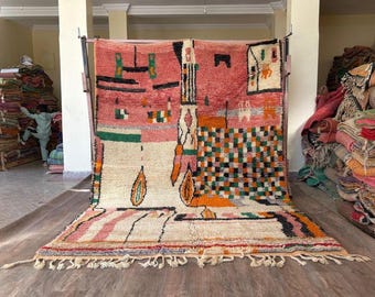 May include: A large, colorful rug with a geometric design. The rug features a mix of colors, including pink, orange, green, and white. The rug has a fringe along the bottom edge and is displayed on a wooden floor.
