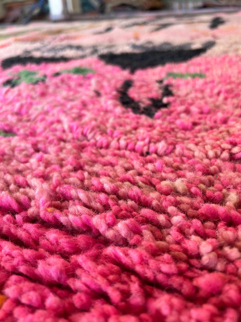 May include: A thick, high-pile rug featuring a vibrant pink base with textured, knotted wool fibers. The surface displays abstract patterns in black and muted green, creating a tactile, shaggy appearance with a deep, dense, and soft pile construction.