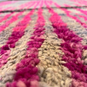 May include: Close-up of a textured rug with a pattern of vertical stripes. The stripes alternate between vibrant pink and neutral beige, creating a bold contrast. The rug's thick pile suggests a soft, comfortable texture, ideal for home decor.