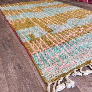 May include: A rectangular rug with a geometric pattern in shades of olive green, light blue, and pink. The rug has a fringe on one end and is made of a textured material. The design features a grid-like pattern with various shapes.