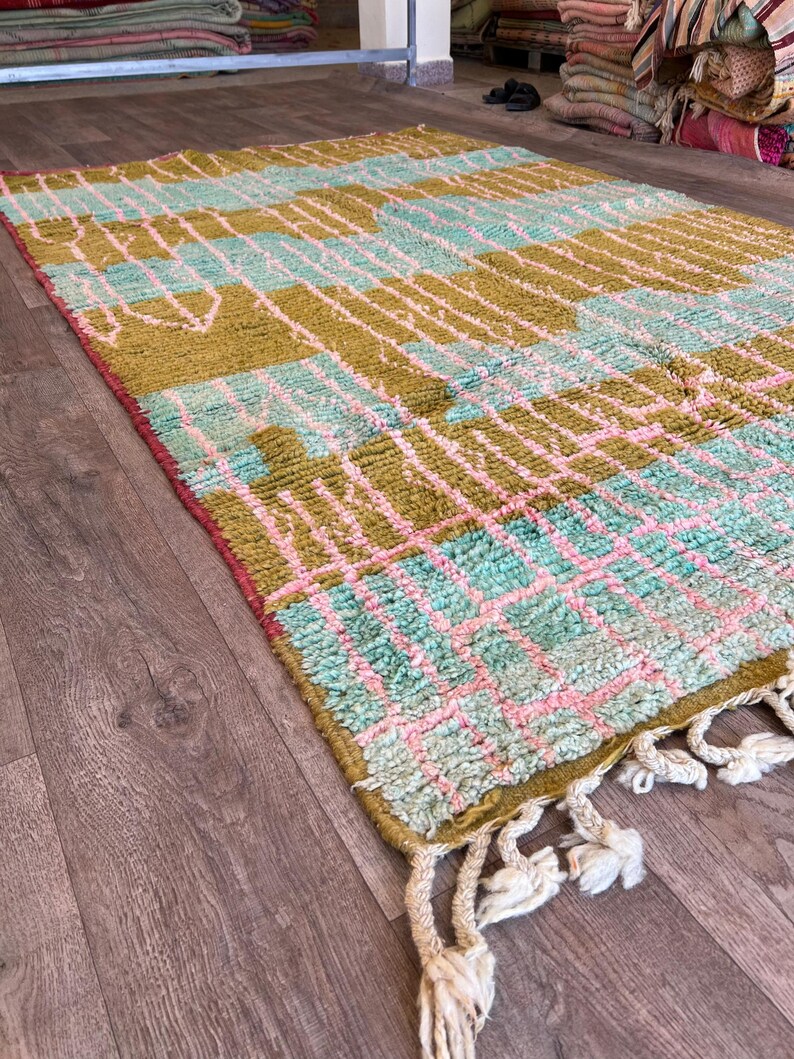 May include: A rectangular rug with a geometric pattern in shades of olive green, light blue, and pink. The rug has a fringe on one end and is made of a textured material. The design features a grid-like pattern with various shapes.