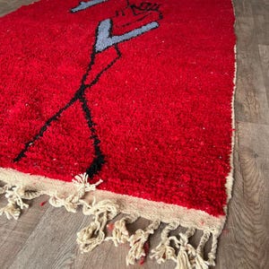 Peut inclure: Un tapis rectangulaire &agrave; poils longs d'un rouge profond, orn&eacute; d'un motif abstrait stylis&eacute; en noir et gris clair. Le tapis est dot&eacute; d'une bordure couleur cr&egrave;me et d'une &eacute;paisse frange nou&eacute;e de la m&ecirc;me couleur sur le bord inf&eacute;rieur.