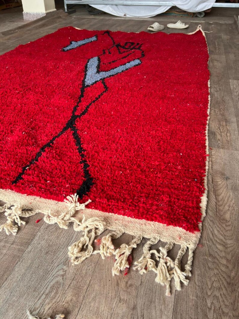 Peut inclure: Un tapis rectangulaire &agrave; poils longs d'un rouge profond, orn&eacute; d'un motif abstrait stylis&eacute; en noir et gris clair. Le tapis est dot&eacute; d'une bordure couleur cr&egrave;me et d'une &eacute;paisse frange nou&eacute;e de la m&ecirc;me couleur sur le bord inf&eacute;rieur.