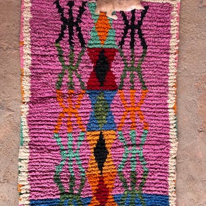 May include: A colourful, hand-woven rug with a geometric pattern. The rug has a pink base with black, orange, green, blue, and red geometric shapes. The edges are finished with a cream-coloured fringe. A small, light-coloured cat is resting on the rug.