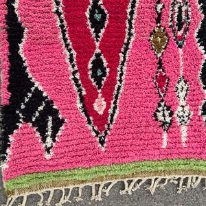 Puede incluir: Una vibrante alfombra marroqu&iacute; rosa y roja con un patr&oacute;n geom&eacute;trico de diamantes. La alfombra presenta detalles en negro y blanco, un borde verde y un borde con flecos. La textura gruesa y afelpada de la alfombra a&ntilde;ade calidez y estilo a cualquier espacio.