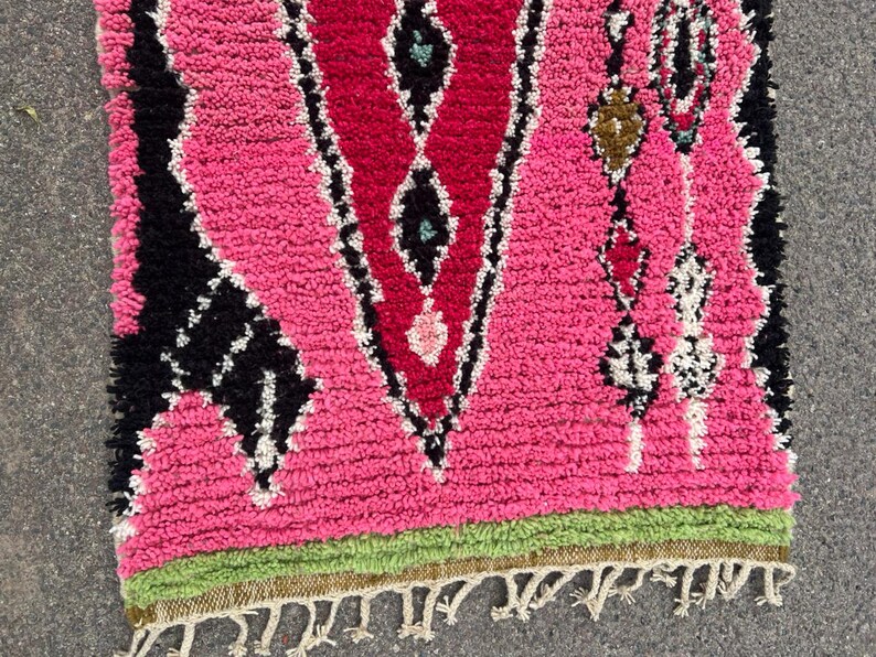 Puede incluir: Una vibrante alfombra marroqu&iacute; rosa y roja con un patr&oacute;n geom&eacute;trico de diamantes. La alfombra presenta detalles en negro y blanco, un borde verde y un borde con flecos. La textura gruesa y afelpada de la alfombra a&ntilde;ade calidez y estilo a cualquier espacio.