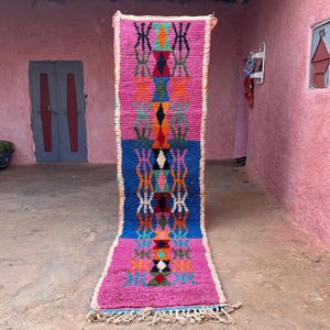 May include: A colourful, rectangular, hand-woven rug with a geometric design. The rug features pink and blue sections with orange, green, and black patterns. It has a fringed edge and is displayed vertically, likely for sale.