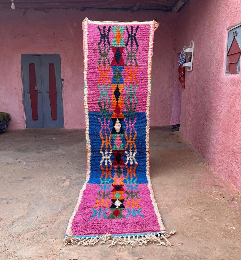 May include: A colourful, rectangular, hand-woven rug with a geometric design. The rug features pink and blue sections with orange, green, and black patterns. It has a fringed edge and is displayed vertically, likely for sale.