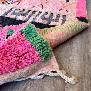 May include: A hand-knotted wool rug featuring a light pink base with vibrant geometric patterns in black, hot pink, and green. The rug has a thick, shaggy pile texture, a folded corner revealing the woven backing, and cream-colored fringe tassels.