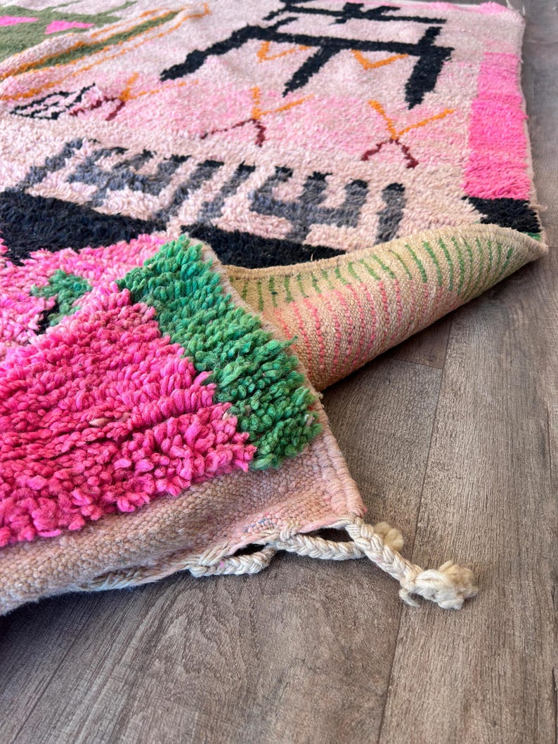 May include: A hand-knotted wool rug featuring a light pink base with vibrant geometric patterns in black, hot pink, and green. The rug has a thick, shaggy pile texture, a folded corner revealing the woven backing, and cream-colored fringe tassels.