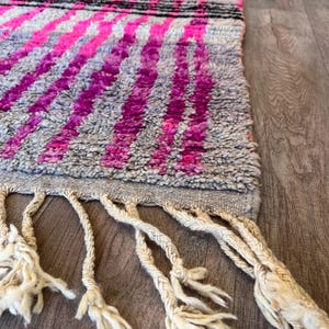 May include: A grey rug featuring a pattern of vibrant pink stripes and a black stripe. The rug has a textured surface and cream-coloured fringe along the bottom edge. The rug is on a wooden surface.