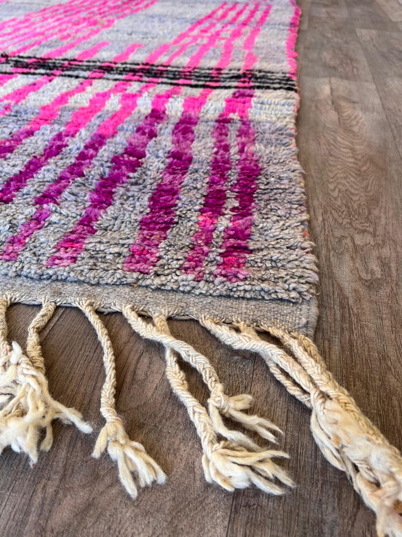 May include: A grey rug featuring a pattern of vibrant pink stripes and a black stripe. The rug has a textured surface and cream-coloured fringe along the bottom edge. The rug is on a wooden surface.