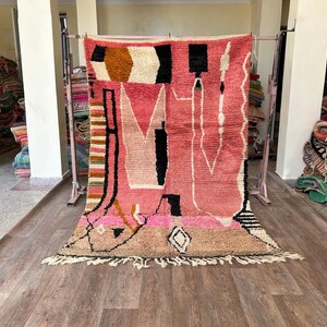 May include: A pink, black, and white Moroccan rug with geometric patterns and fringe. The rug is hanging on a rack in a shop.