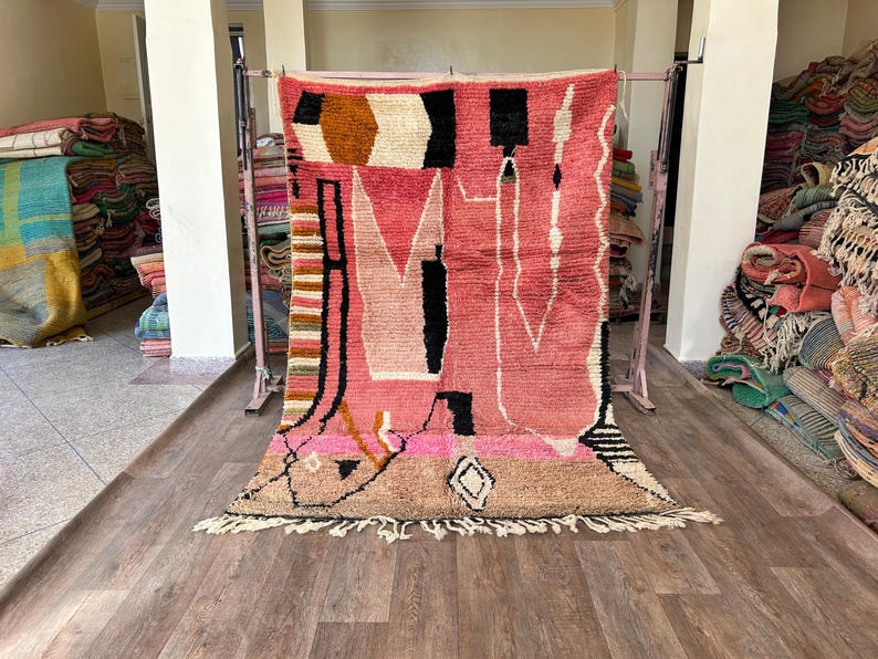 May include: A pink, black, and white Moroccan rug with geometric patterns and fringe. The rug is hanging on a rack in a shop.