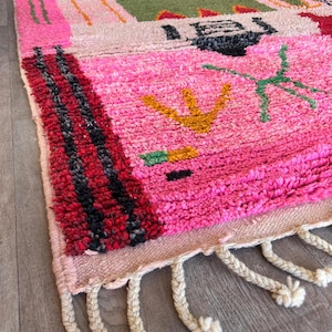 May include: A rectangular area rug featuring a high-pile texture with a vibrant pink base. The design includes abstract geometric shapes in green, black, yellow, and red, with thick black and red striped borders and cream-colored braided fringe.